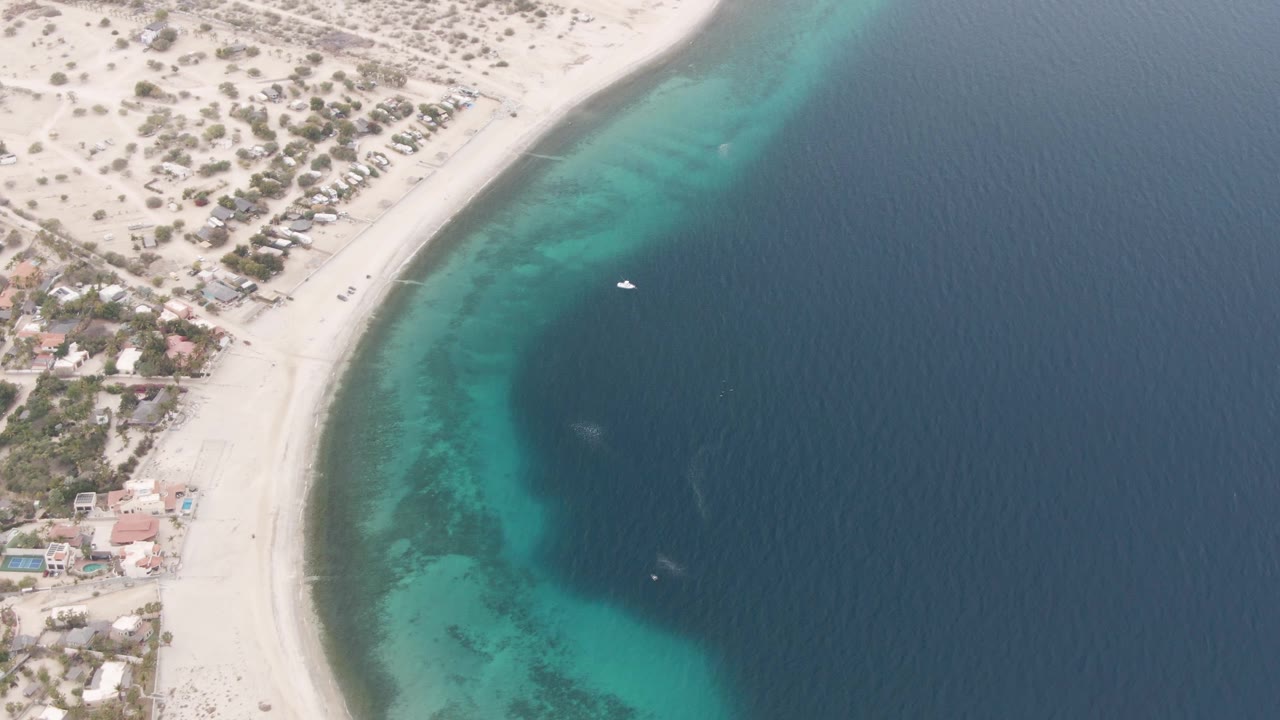 Aerial frame over Los Barriles showing a curved pale beach, turquoise shallows, darker offshore water, nearby coastal buildings, sparse vegetation, and no visible people.