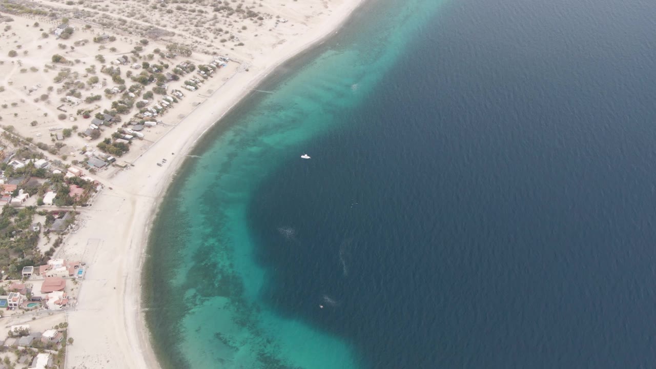 Aerial view in Los Barriles with the small white boat slightly farther across the dark water, turquoise shallows lining the beach, coastal buildings at lower left, and no visible people.