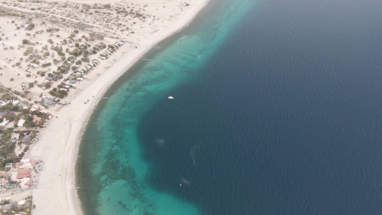 Overhead Los Barriles shoreline frame showing the beach arc tighter along the left edge, turquoise water near shore, a small boat in open water, land vegetation, buildings, and no visible people.