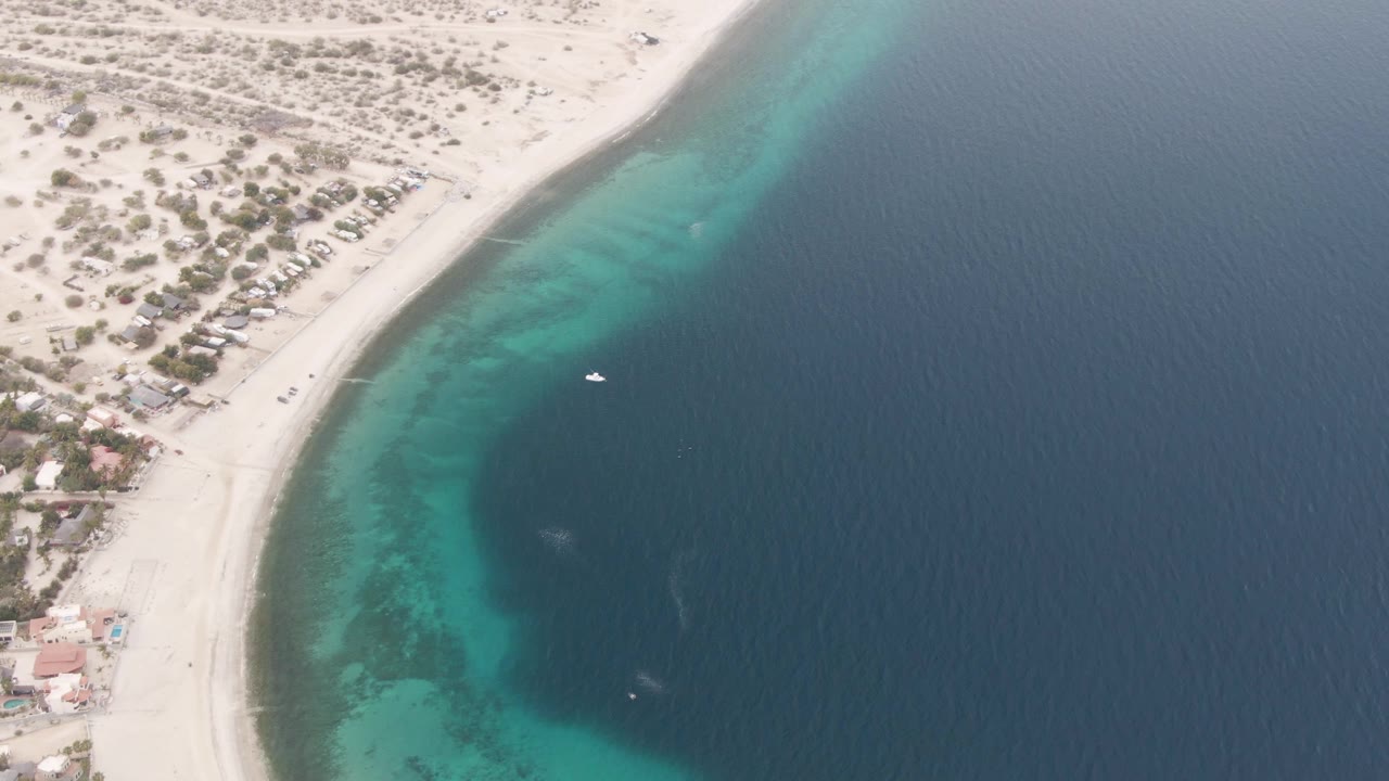 Aerial coastline frame with pale sand, turquoise shallows, and a boat offshore.