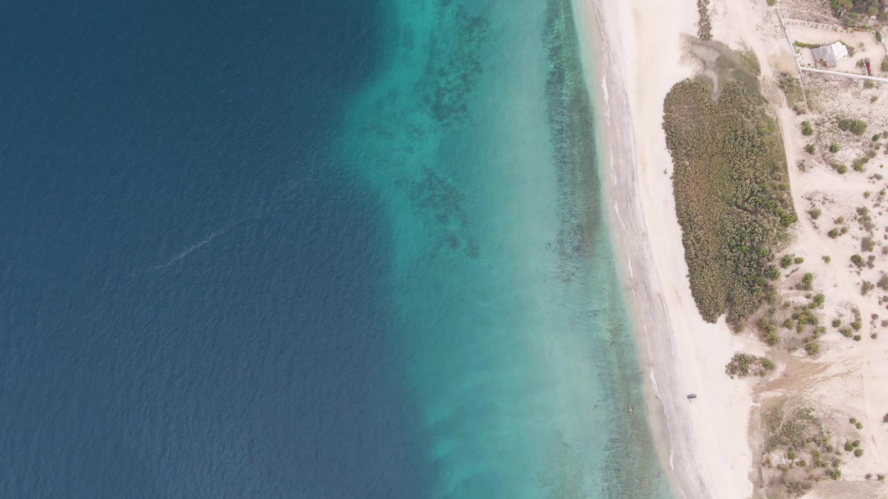Aerial frame over Los Barriles showing a faint boat wake in the darker offshore water, turquoise shallows beside the beach, dune vegetation, and no visible people.