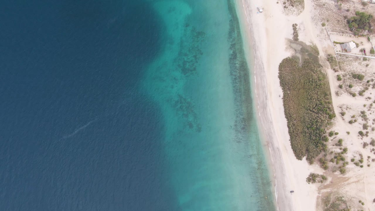 Top-down aerial view in Los Barriles, Baja California Sur, with deep blue water on the left, turquoise shallows near the sand, a beach road on the right, and no visible people.