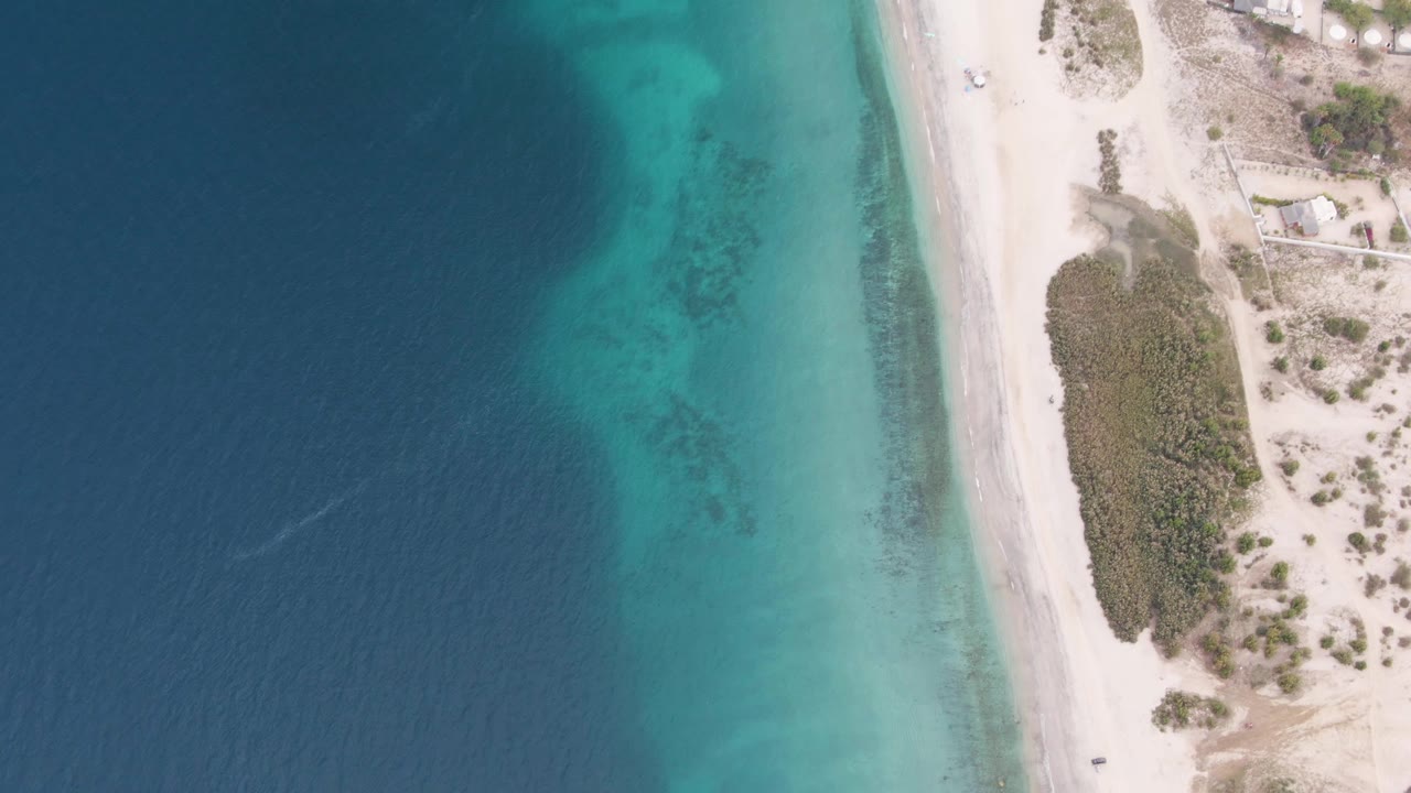 Top-down Los Barriles coastal view with the ocean filling the left half, a narrow turquoise shallows band, pale beach road, sparse vegetation, small structures, and no visible people.