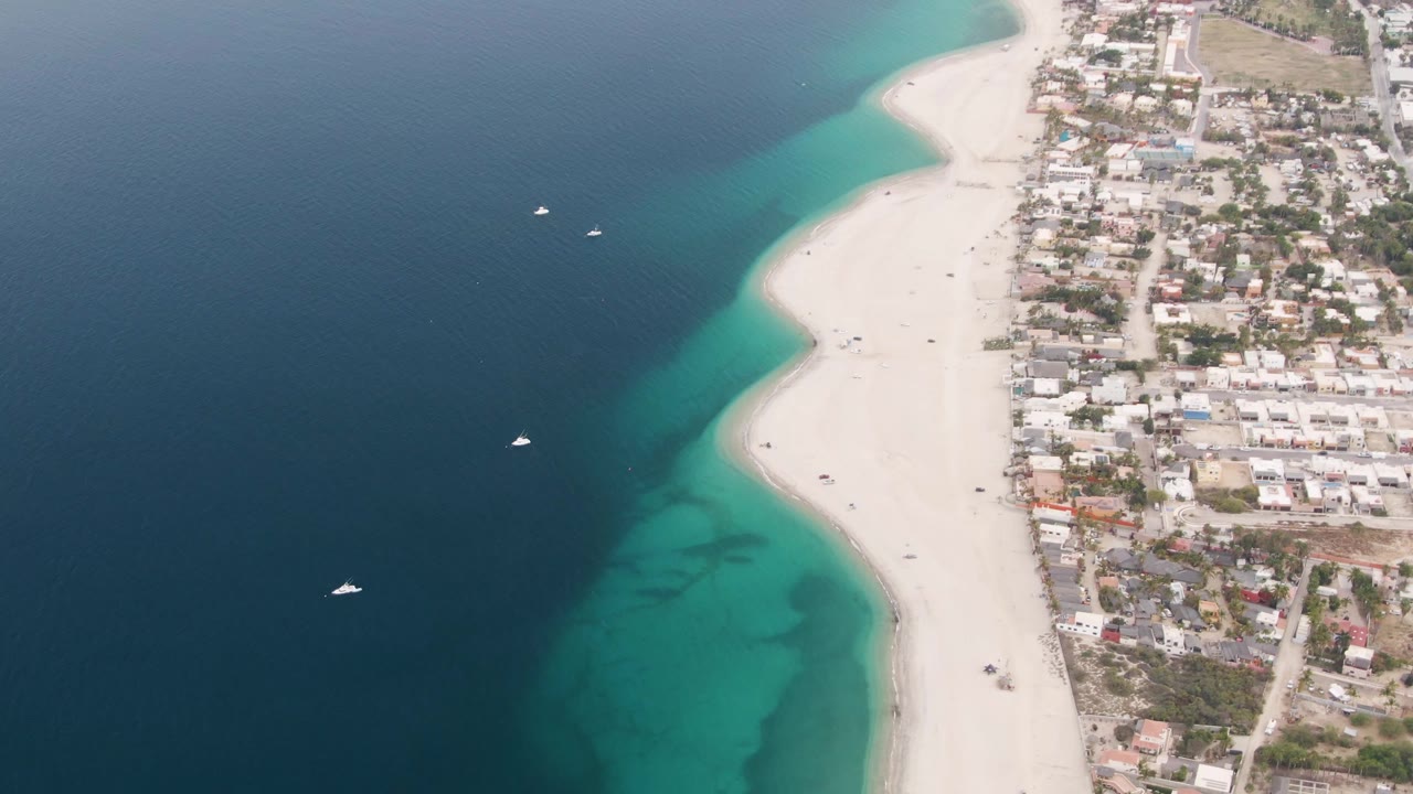 Opening aerial frame over Los Barriles shows deep blue water on the left, turquoise shallows along the sand, a coastal town on the right, and no visible people.