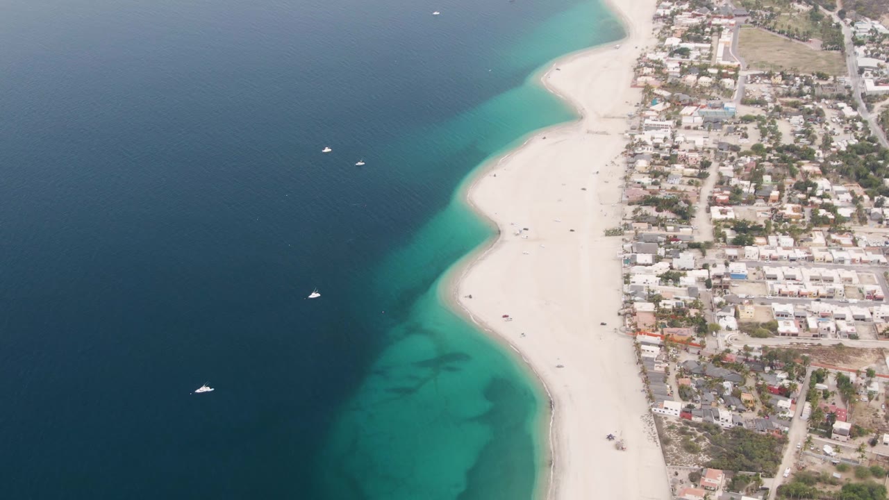 Aerial frame at 0:01 shows the Los Barriles beach curving beside turquoise water, with compact residential blocks and roads filling the right side and no visible people.