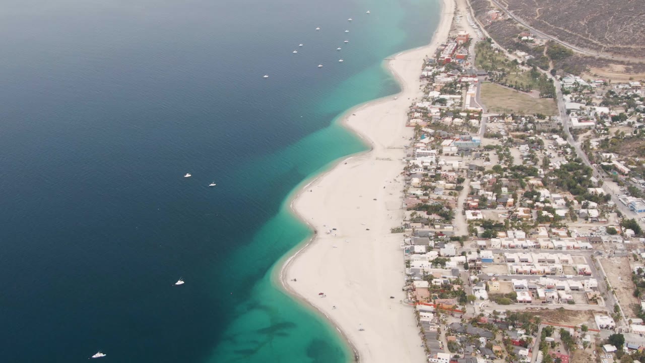 Aerial frame over Los Barriles showing dark blue ocean at left, turquoise shallows along a wide sandy beach, small boats offshore, residential blocks to the right, and no visible people.