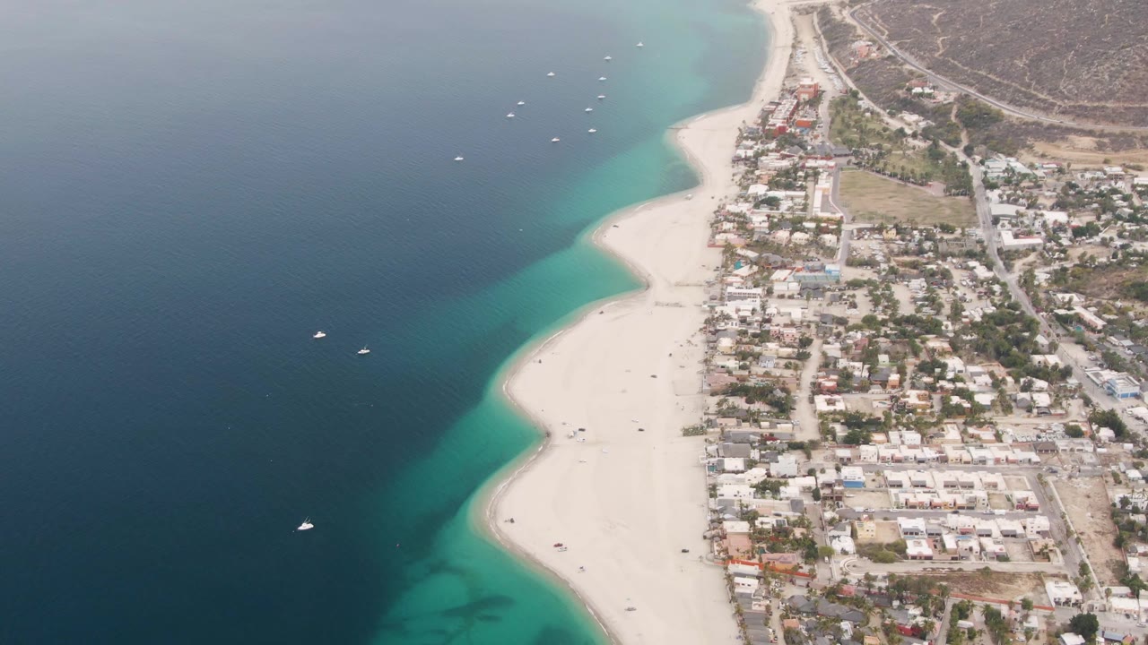 Aerial frame over Los Barriles with more of the pale beach centered, boats spaced across the blue water, clustered homes beside the shore, and no visible people.