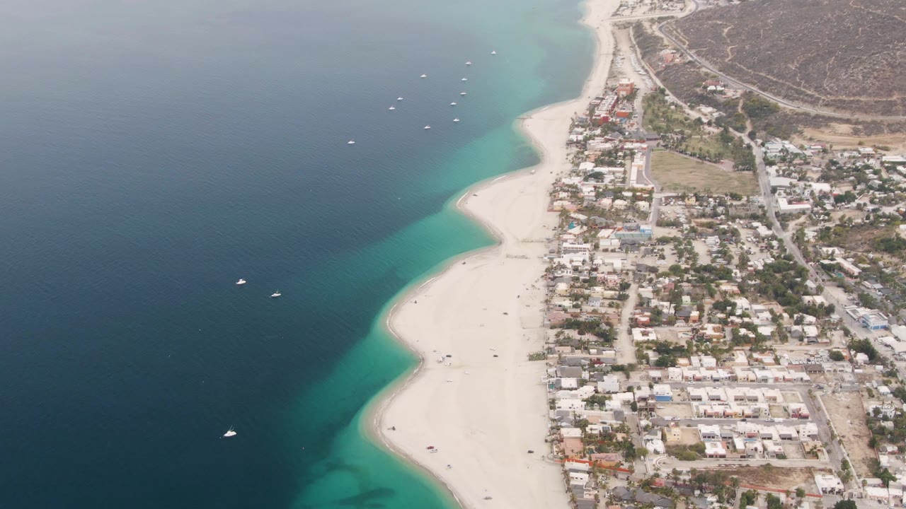 Aerial frame over Los Barriles showing the beach curve, several anchored boats in the water, compact residential buildings inland, dry hills beyond, and no visible people.