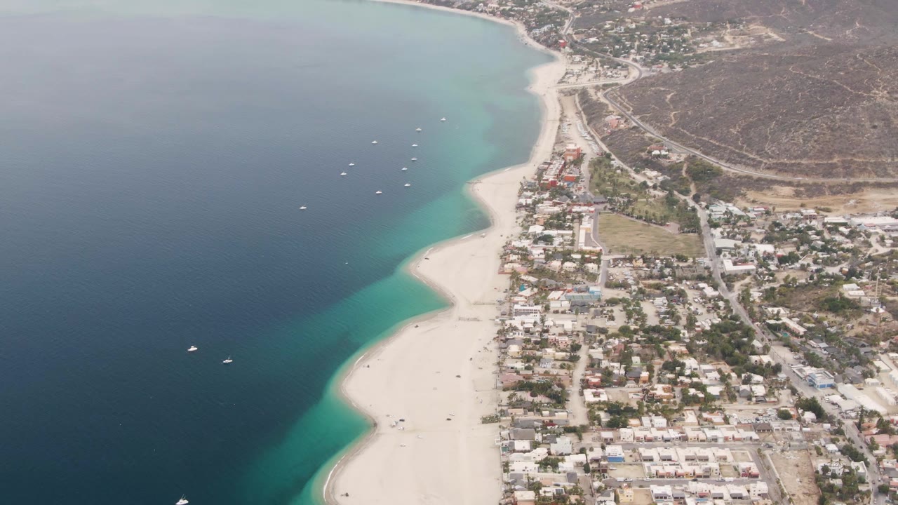 Aerial frame over Los Barriles, Mexico, showing deep blue ocean, turquoise shallows, a curving sandy beach, town buildings, dry hillside terrain, and no visible people.
