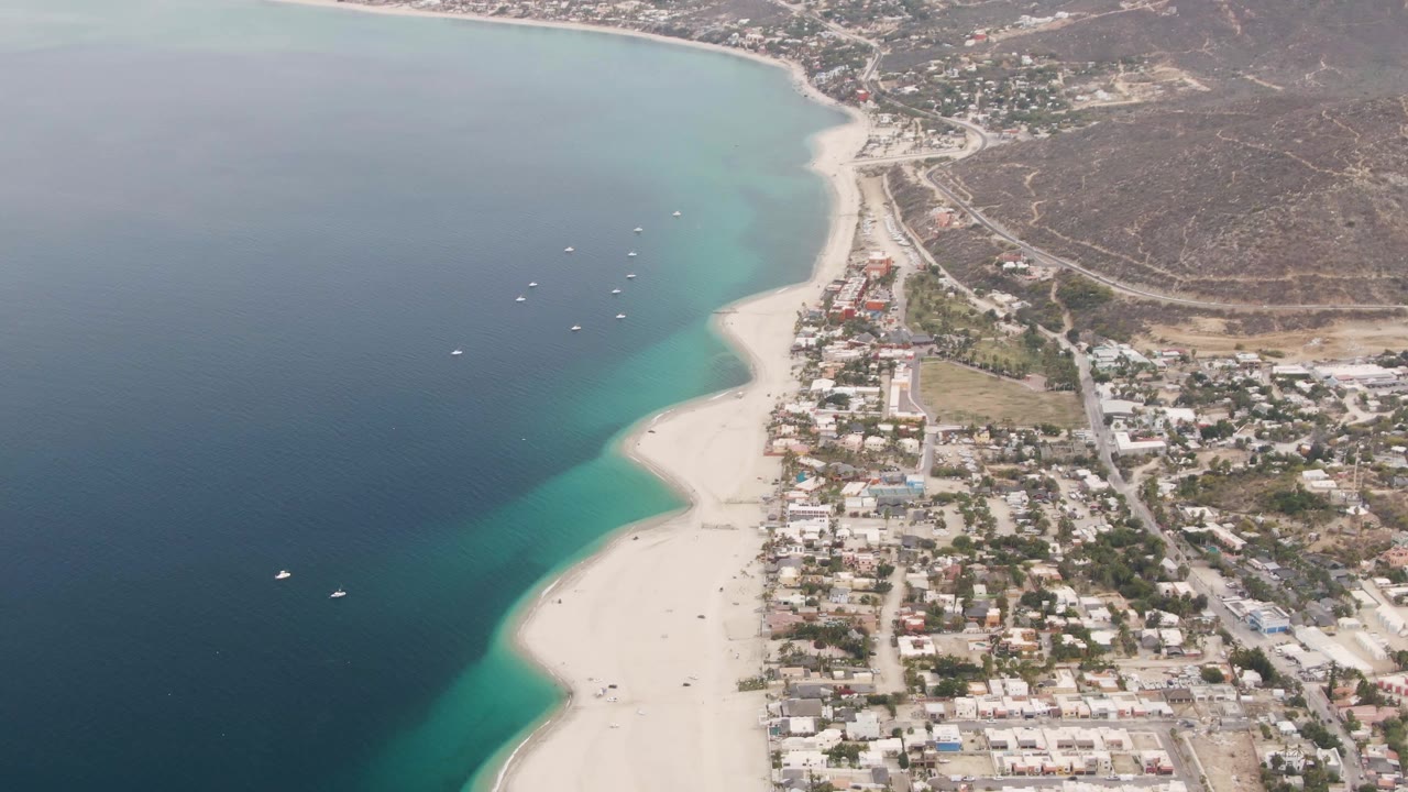 Aerial view in Los Barriles, Mexico, with the ocean on the left, small boats offshore, pale beach at center, town streets to the right, hillside in back, and no visible people.
