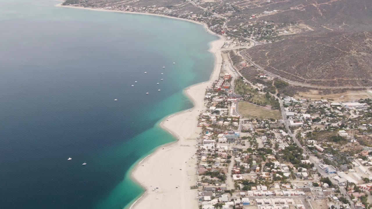 High aerial frame of Los Barriles coastline with turquoise water bands along the sand, deep blue sea farther out, compact buildings and roads inland, dry hills, and no visible people.