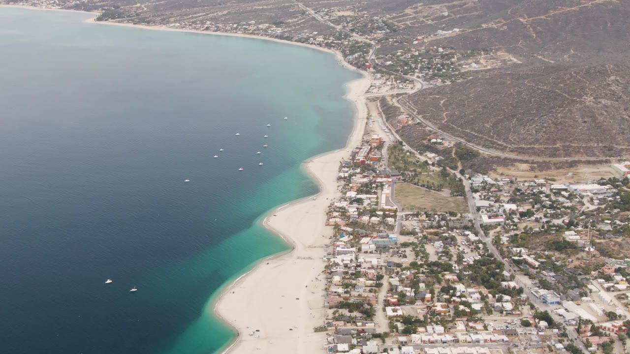 Aerial coastline with ocean, anchored boats, beach, town blocks, and hillside.