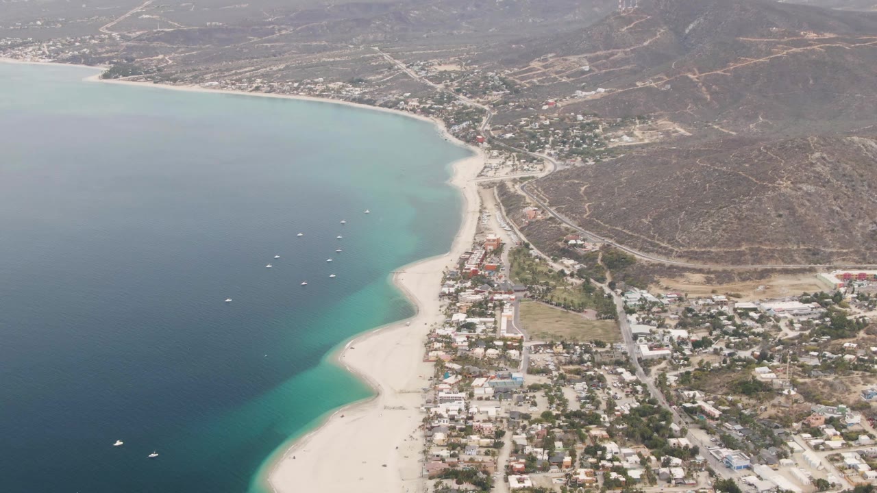 Aerial frame over Los Barriles showing deep blue and turquoise water, a pale sandy beach, clustered town buildings, dry hills, and no visible people.