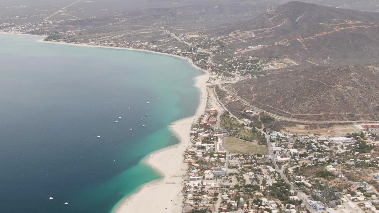 Los Barriles aerial view with several small boats offshore, clear turquoise shallows along the beach, town streets and buildings inland, and no visible people.