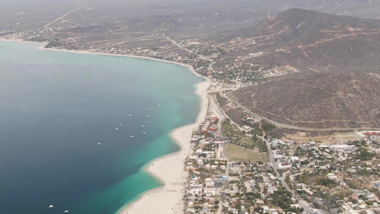 Aerial still from Los Barriles with the shoreline bending across frame, a smaller cluster of visible boats, pale beach sand, hillside roads, and no visible people.