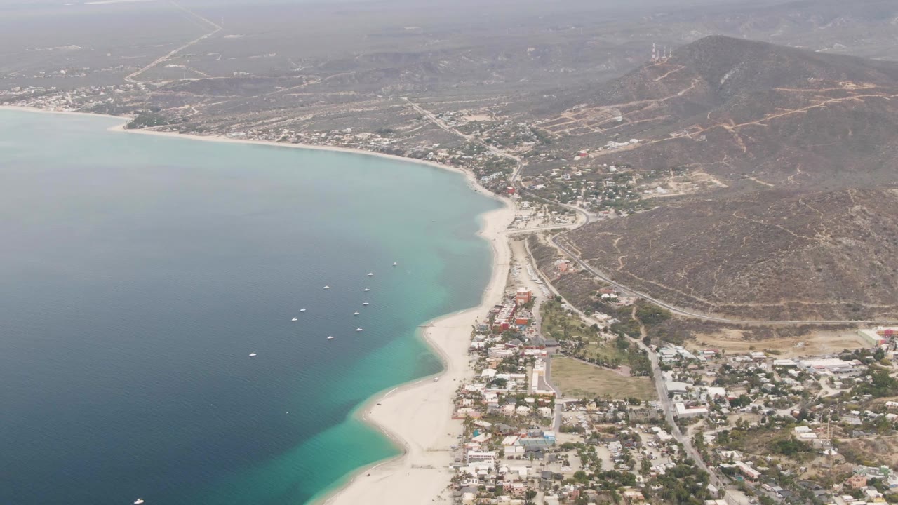 Aerial bay view with beach edge, boats, town buildings, roads, and hills.
