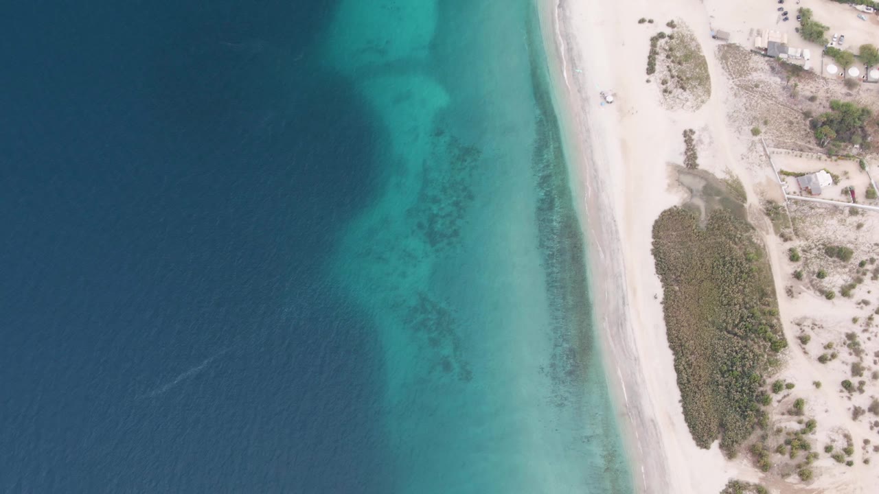 Overhead view in Los Barriles, Baja California Sur, showing dark blue water, turquoise shallows, white sand, dune vegetation, a small white vehicle near the beach road, and no visible people.