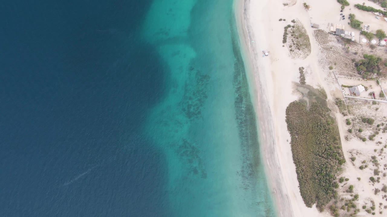Top-down view in Los Barriles with darker offshore water, bright turquoise shallows, a narrow white beach, scrub-covered dunes, small buildings near the upper right, and no visible people.