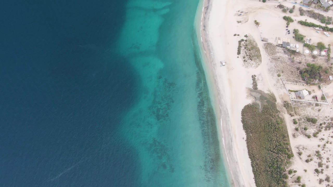 Aerial frame over Los Barriles, Baja California Sur showing deep blue water, turquoise shallows, white sand, scrubby beach vegetation, and no visible people.