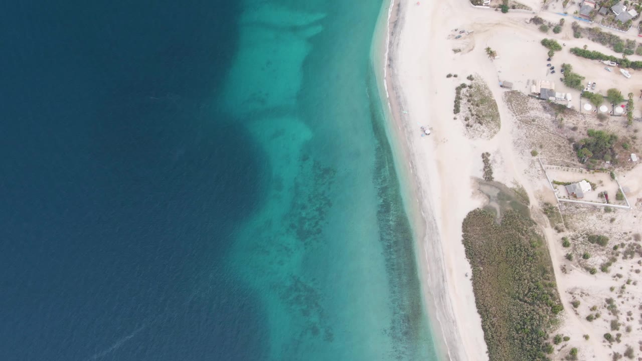 Aerial frame over Los Barriles with a small vehicle on the pale beach, turquoise water along the shore, sparse vegetation inland, and no visible people.