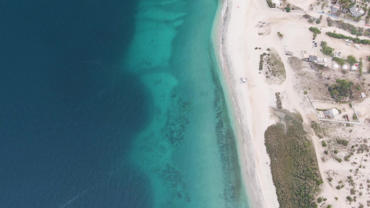 Aerial frame over Los Barriles showing the shoreline shifted slightly through frame, with dark offshore water, bright turquoise shallows, beach structures, scrub, and no visible people.