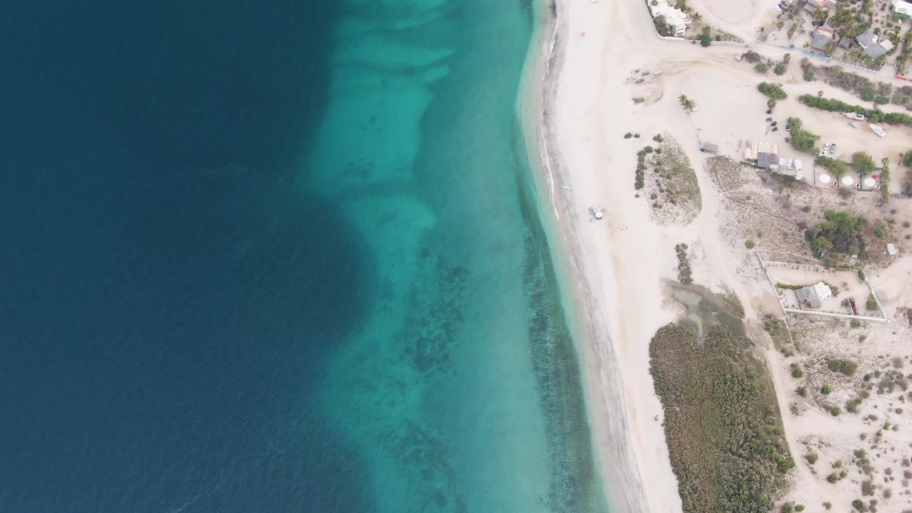 Aerial beach view with clear water bands, white sand, scrub vegetation, and a small vehicle on shore.