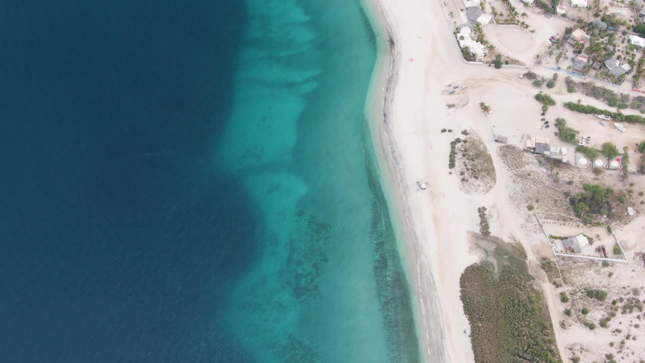 Overhead view of the Los Barriles shoreline with deep blue water on the left, turquoise shallows near the beach, pale sand, scattered buildings, scrub vegetation, and no visible people.