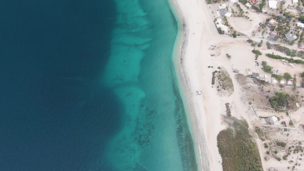 High overhead frame from Los Barriles showing the shoreline slightly farther along, with darker offshore water, bright turquoise patches, white sand, coastal lots, and no visible people.