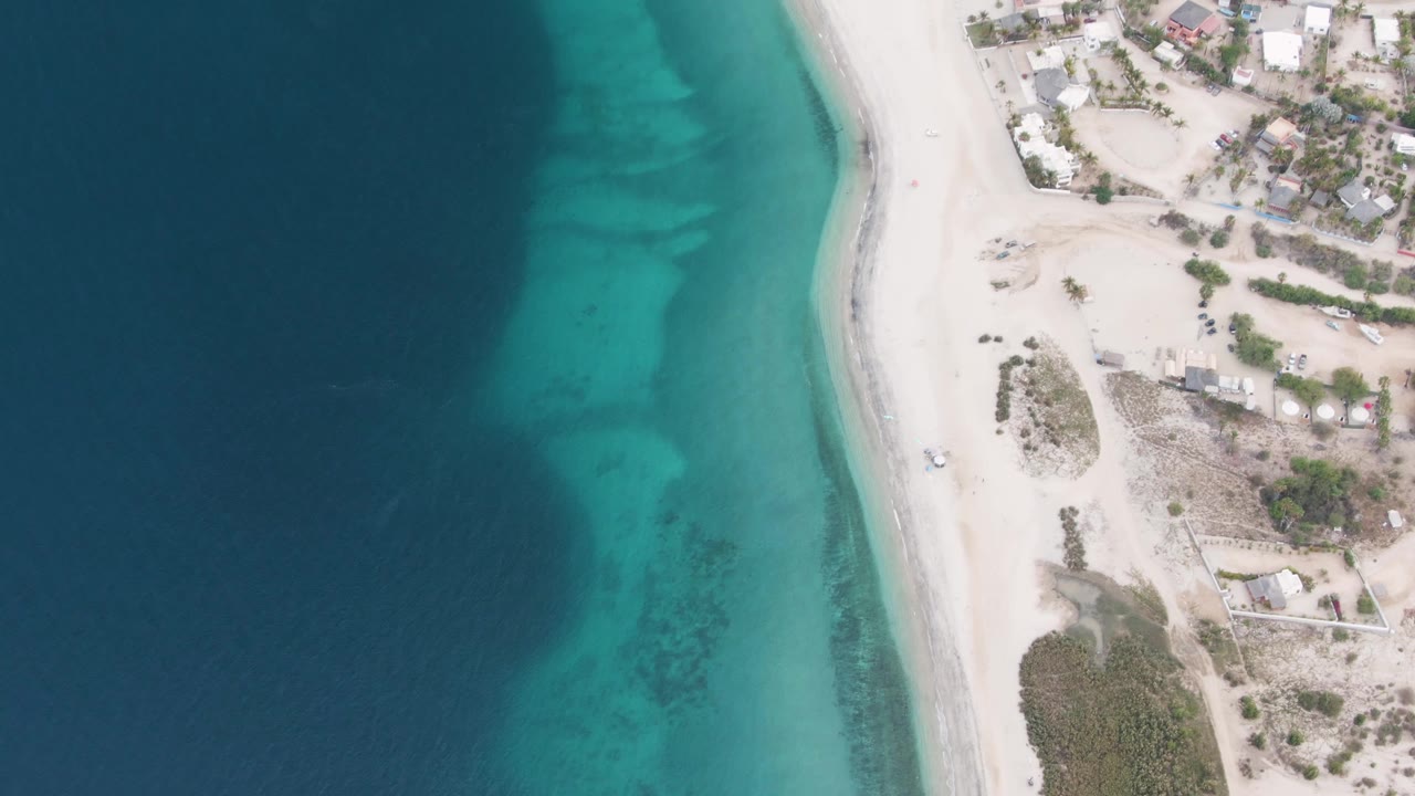 Los Barriles aerial view with a curved band of turquoise water against pale sand, dry vegetation and buildings on the land side, and no visible people on the beach.