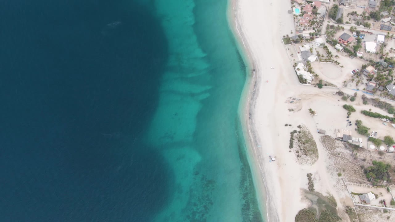 Opening overhead frame of Los Barriles shoreline with deep blue water fading into turquoise shallows, pale sand, beachside buildings, sparse vegetation, and no visible people.