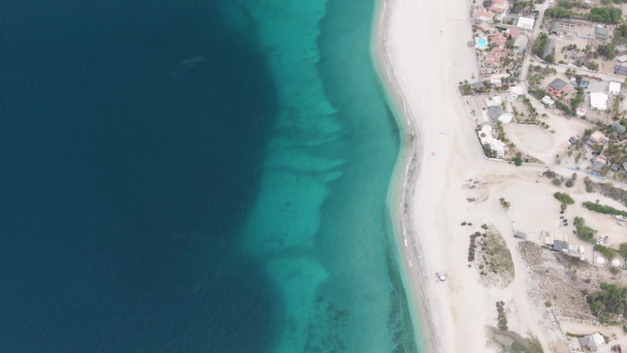 Third frame over Los Barriles with dark ocean occupying the left half, a bright turquoise band near shore, pale beach sand, scattered houses, and no visible people.