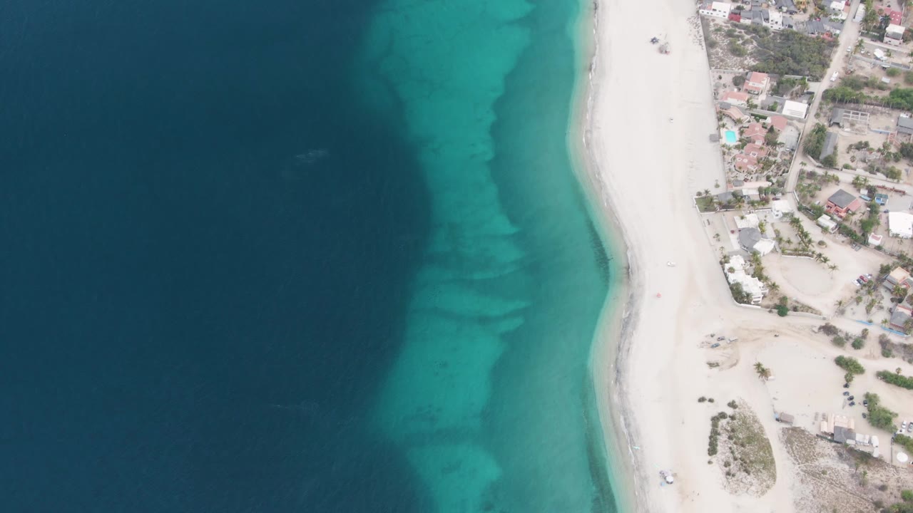 Overhead frame of Los Barriles showing dark blue ocean fading into turquoise shallows beside a white sand beach and shoreline buildings, with no visible people.