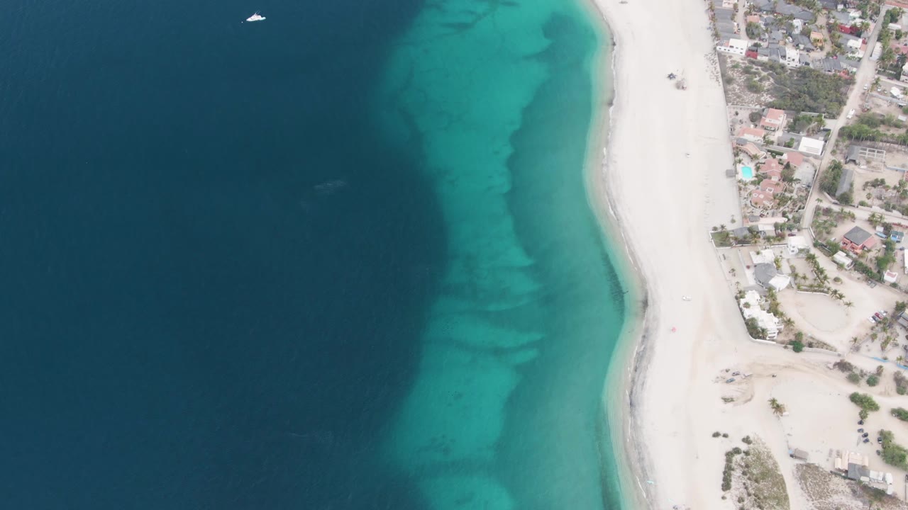 A small white boat appears near the top of the Los Barriles ocean frame, above turquoise shallows, white sand, and beachfront buildings, with no visible people.