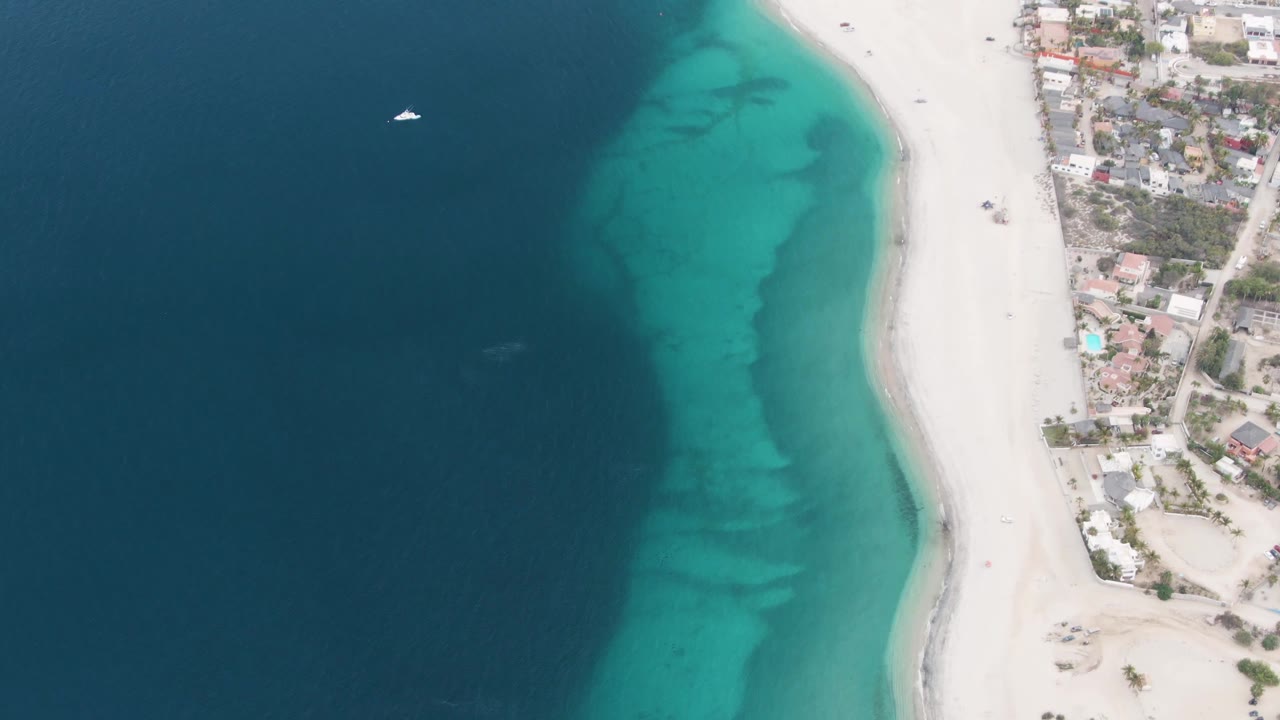 High overhead view in Los Barriles, Baja California Sur with dark blue ocean on the left, turquoise shallows near the sand, coastal buildings at the right edge, a small sailboat offshore, and no visible people.