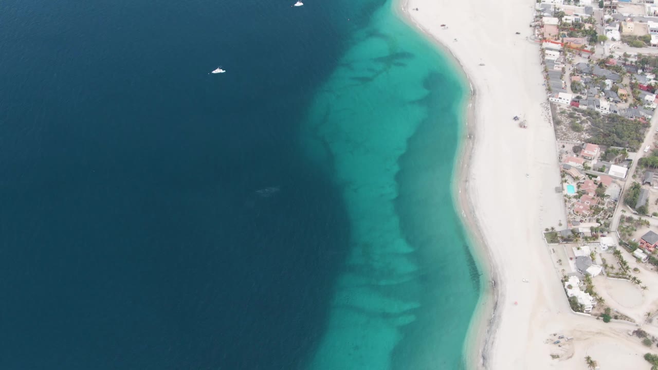 Overhead coastline with a sailboat offshore, turquoise water over seabed, white sand, and buildings along shore.
