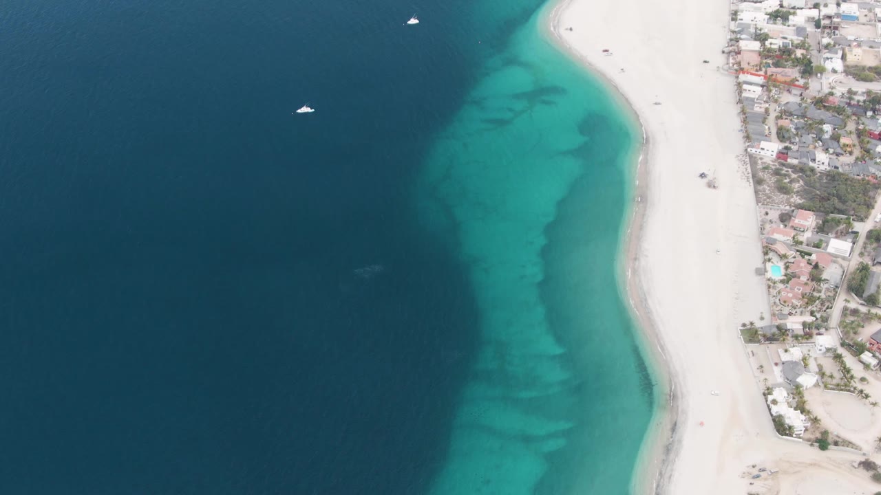 Los Barriles aerial frame with the beach and buildings pushed toward the right side, darker open water taking most of the left side, a small white sailboat near the upper water, and no visible people.