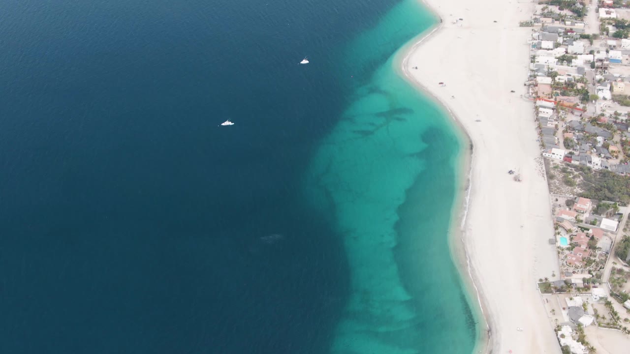 Overhead aerial frame of Los Barriles shoreline with deep blue water, turquoise shallows, white sand, small boats offshore, beachside buildings, and no visible people.