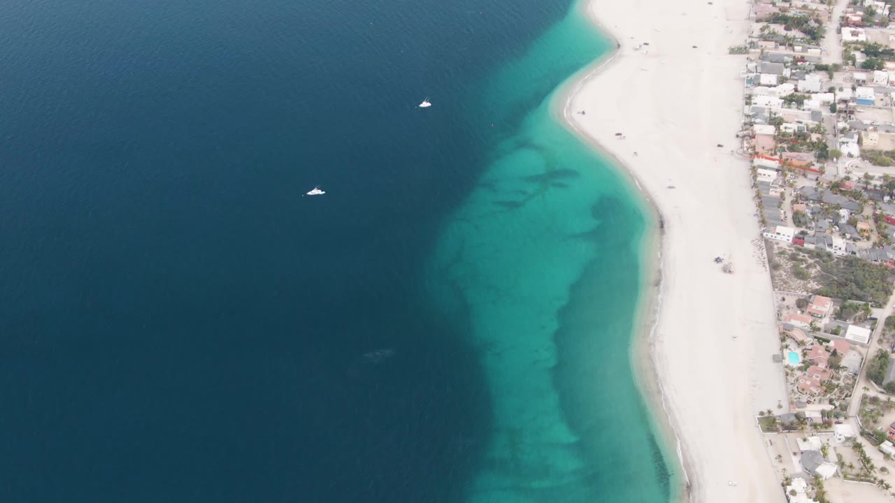 Drone view of clear shallow water beside a curving beach and coastal buildings.