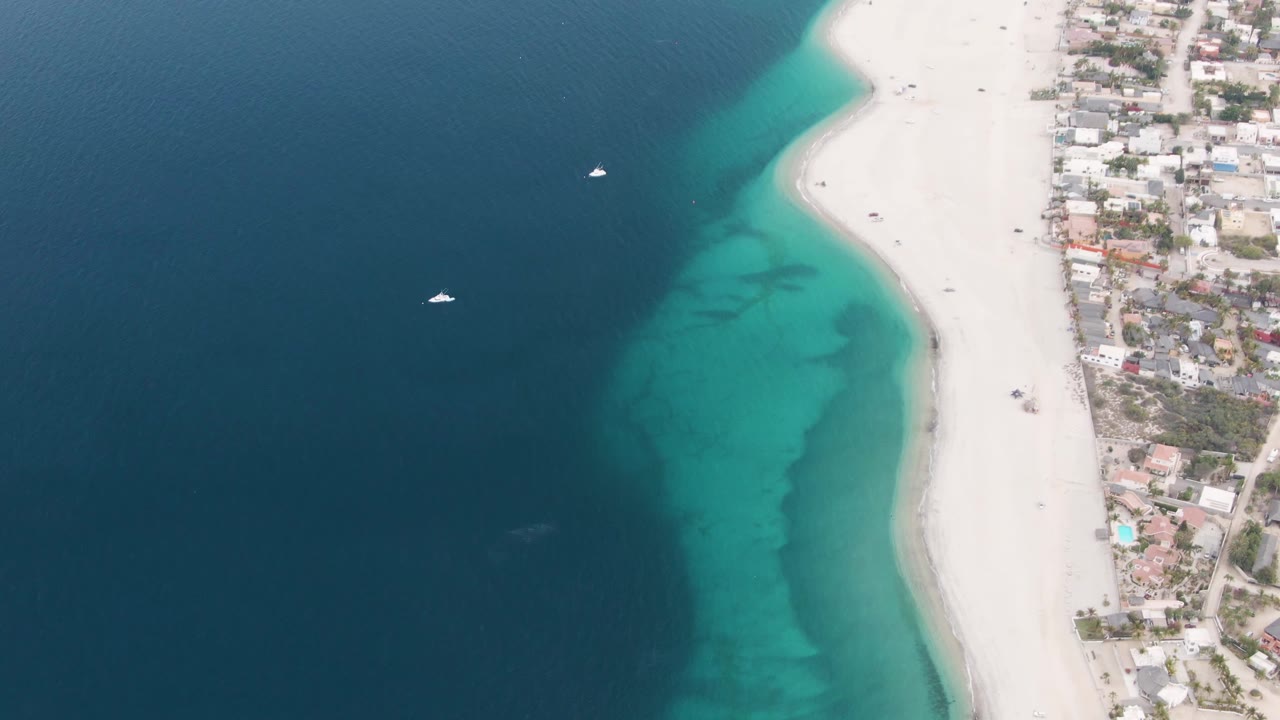 Los Barriles aerial frame with dark open ocean filling the left side, a turquoise nearshore band, white beach on the right, clustered coastal buildings, and no visible people.