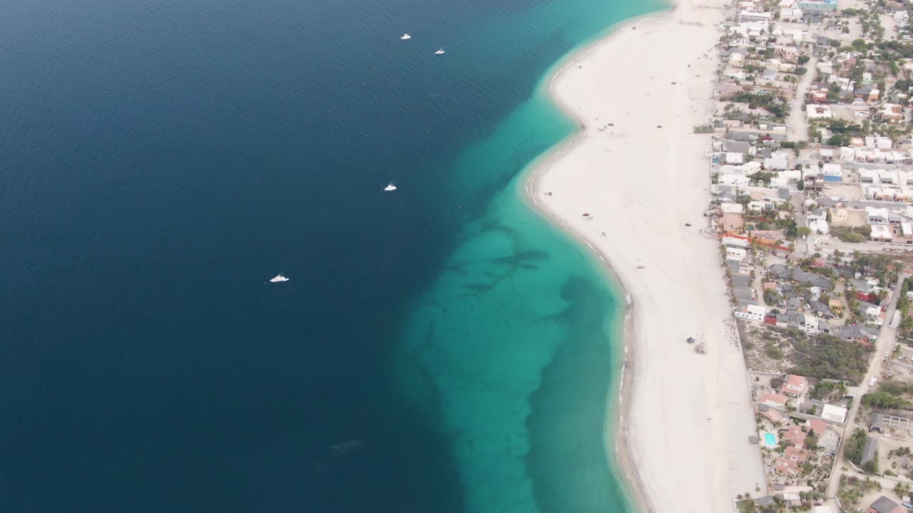 High aerial frame over Los Barriles, Mexico, with dark blue ocean at left, turquoise water along a white sand beach, coastal buildings at right, small offshore boats, and no visible people.