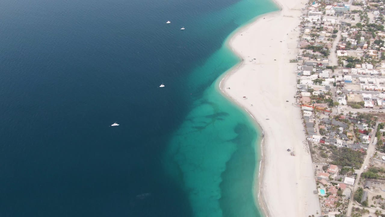 Aerial view in Los Barriles showing a curved white beach between dark ocean and turquoise shallows, with boats offshore, compact town blocks to the right, and no visible people.