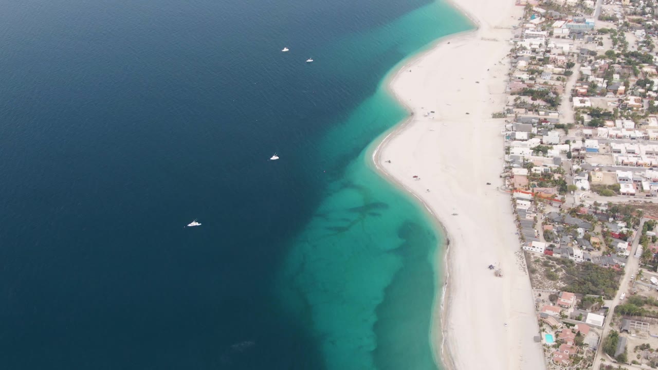 Overhead Los Barriles frame with the beach running vertically through the image, deep water on the left, blue-green shallows near shore, clustered buildings on the right, boats offshore, and no visible people.