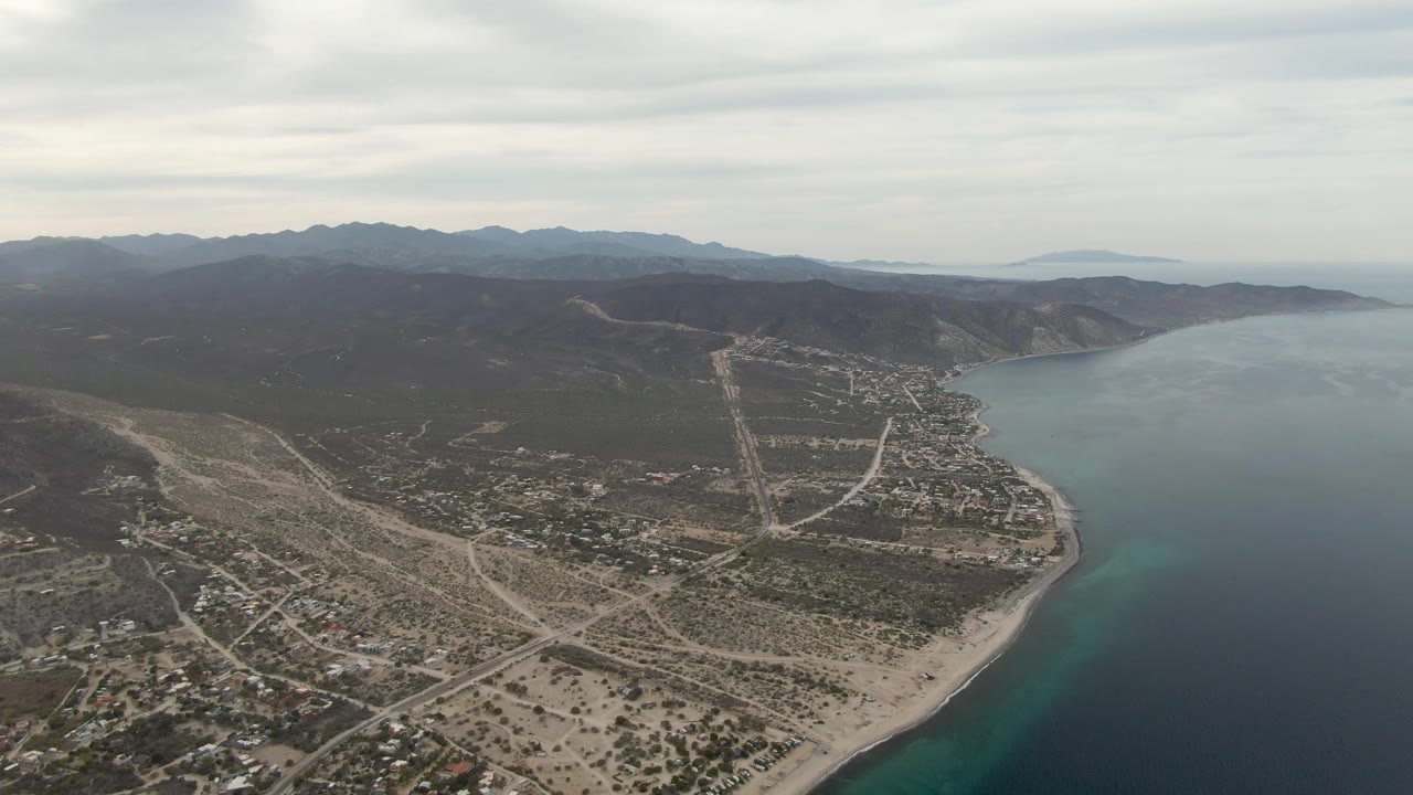 Aerial opening frame over Los Barriles, Baja California Sur, showing sandy shoreline, dark ocean water, dry roads, scattered buildings, distant mountains, and no visible people.
