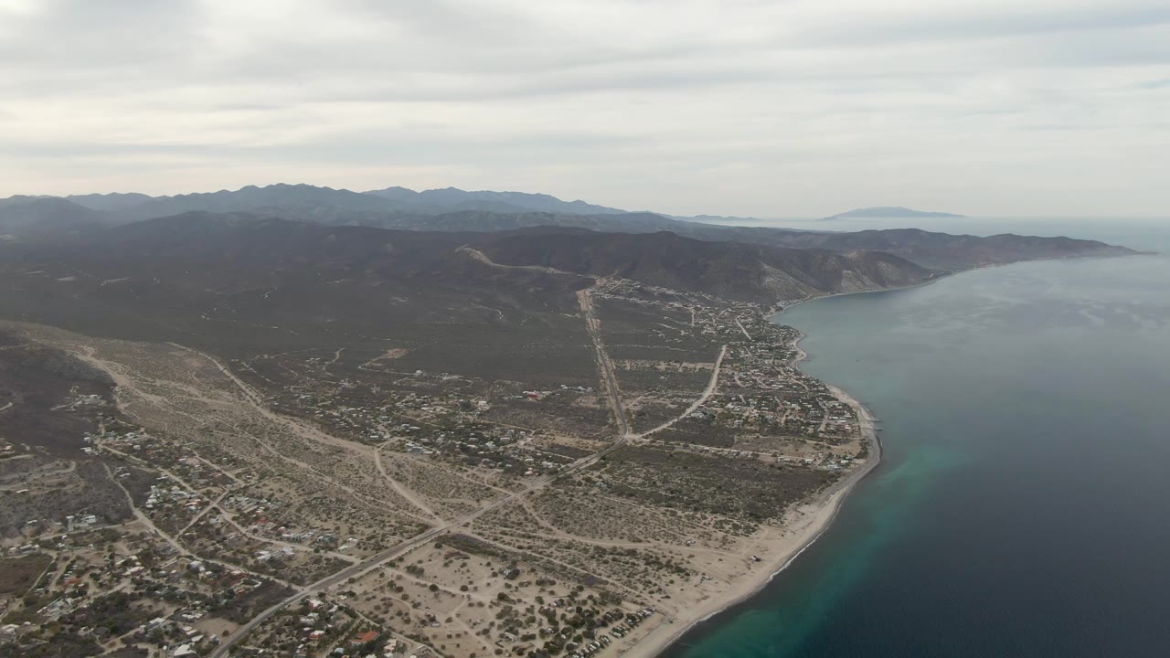 Aerial view over Los Barriles with the road grid and dry lots more centered, the sandy beach curving along the ocean, mountains beyond, and no visible people.