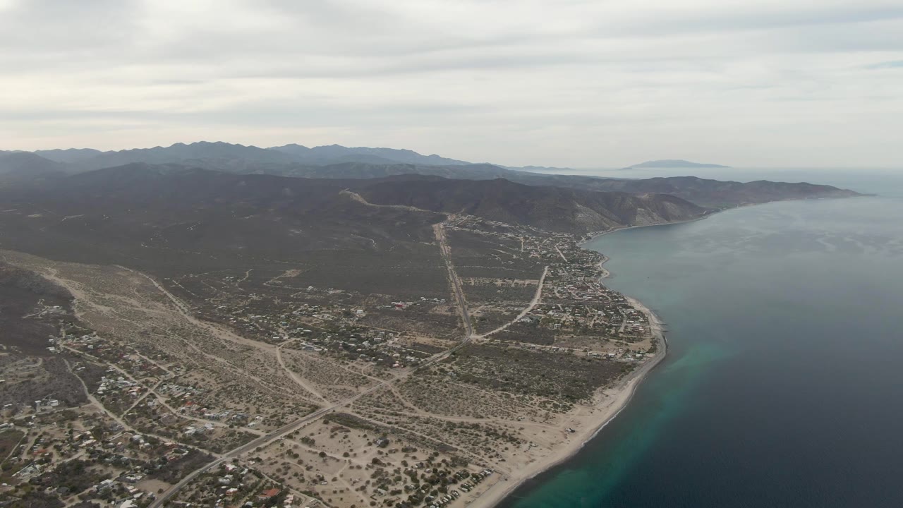 Aerial frame of Los Barriles showing the coastline pushed toward the right edge, a broad spread of roads and buildings on arid ground, mountain ridges, and no visible people.