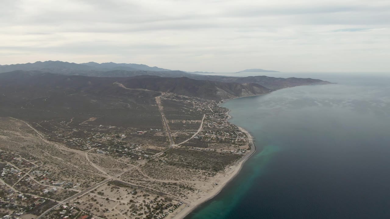 Aerial frame over Los Barriles with the coastline running diagonally, dark blue ocean to the right, pale roads and buildings inland, mountain ridges in the background, and no visible people.