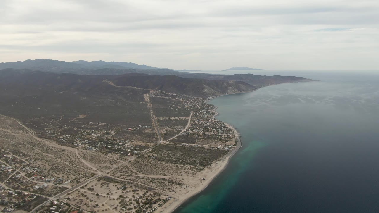 Wide drone view of Los Barriles with sandy beach below, a strip of turquoise water near shore, darker open ocean, inland roads, low buildings, mountains, and no visible people.