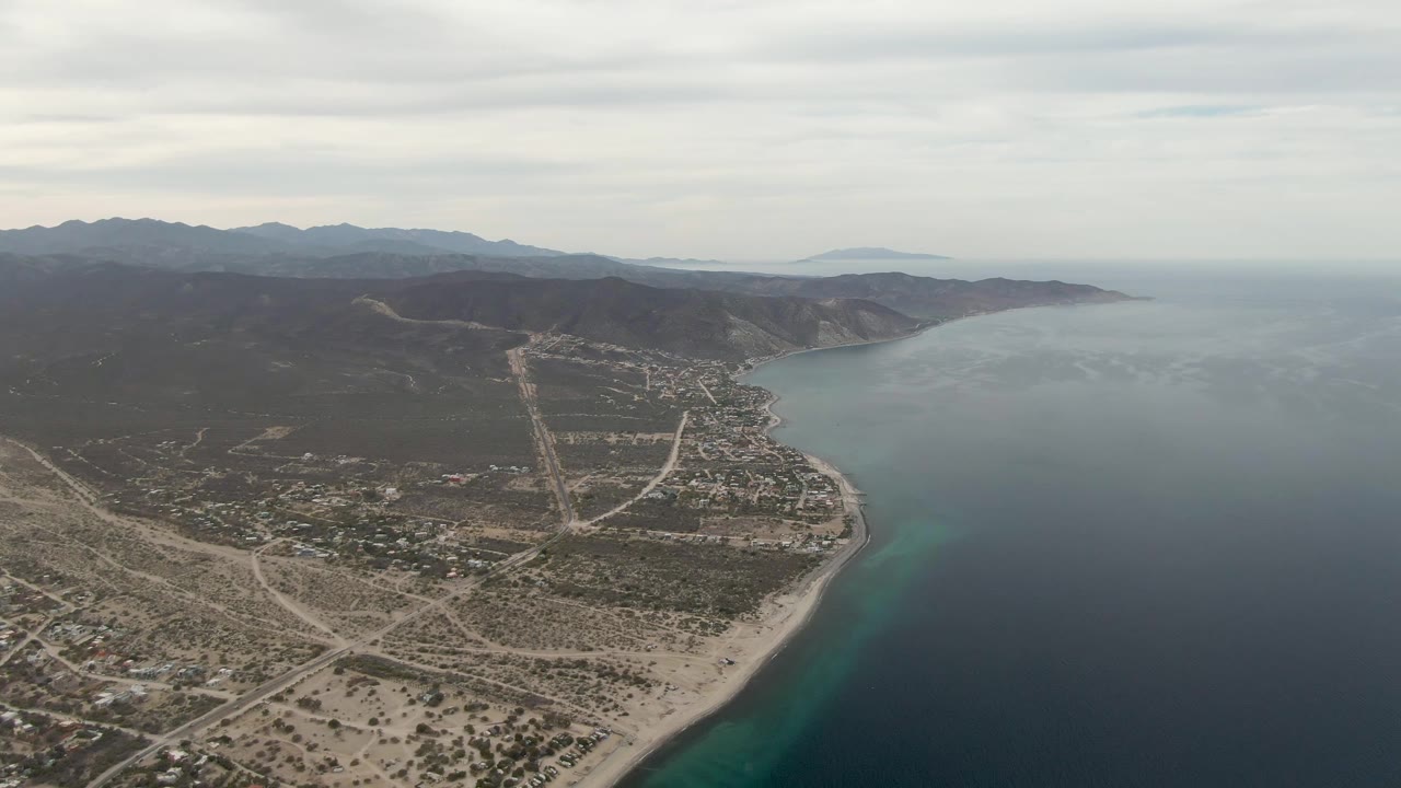 Later aerial frame of Los Barriles showing more of the inland sandy road network, scattered coastal buildings, beach curve, calm ocean surface, mountain backdrop, and no visible people.