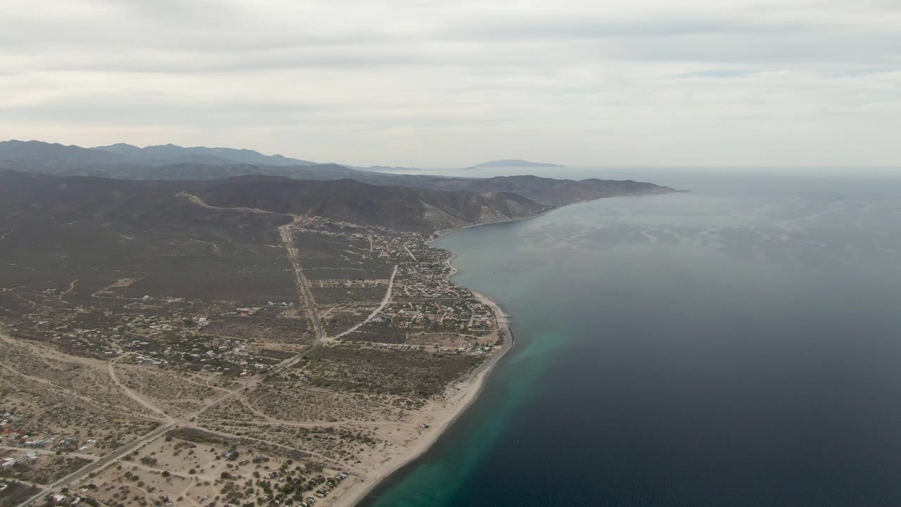 Aerial view of a sandy coastal town beside blue ocean and mountains.