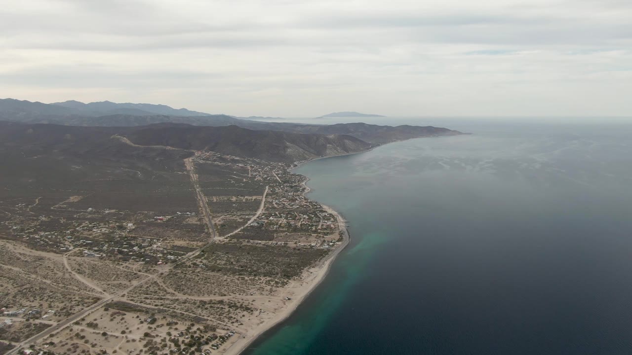 Aerial frame over Los Barriles showing the shoreline running diagonally, pale roads through the settlement, arid land in the foreground, blue ocean on the right, and no visible people.